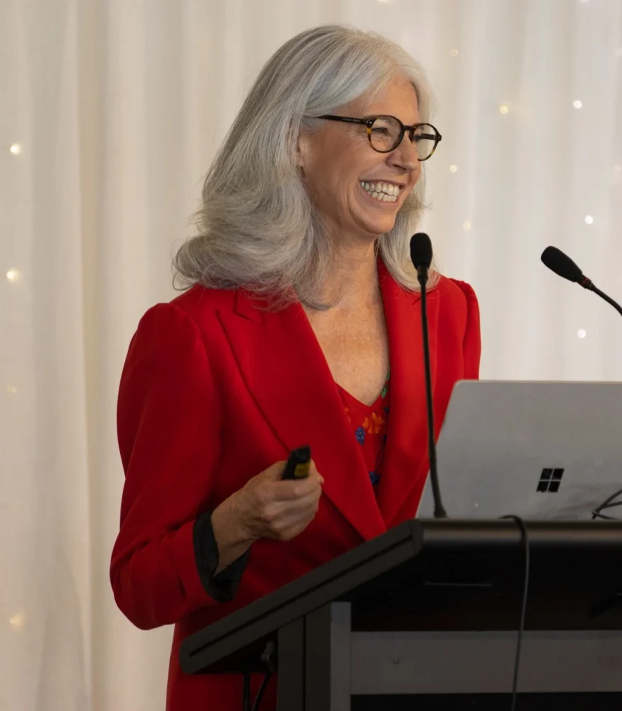 Dr Mary Birdsall standing behind a podium and a laptop, addressing an audience at a seminar